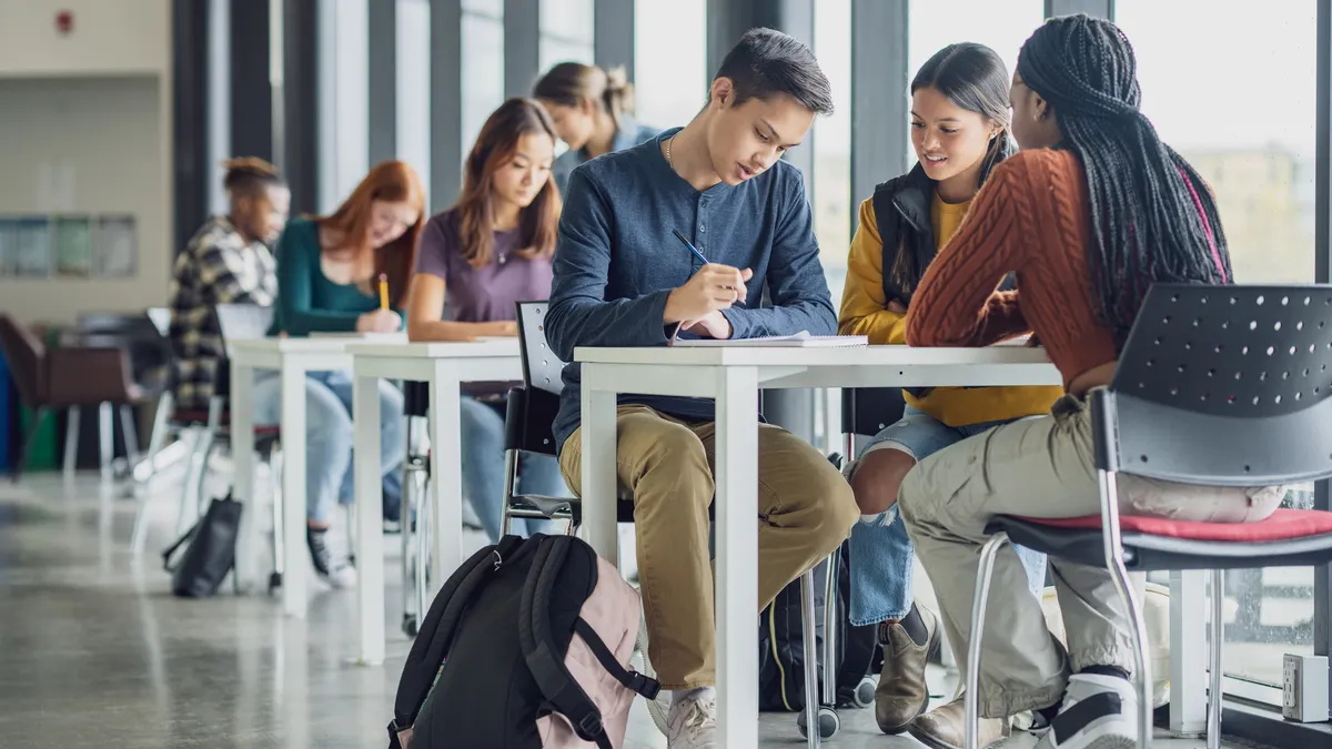 Students are sitting at several tables working in groups. There are laptops and notebooks on the tables and the tables are pushed to a wall of windows.