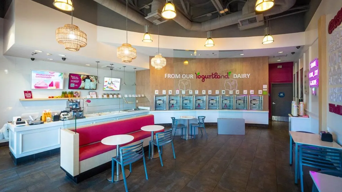 An image of the inside of a Yogurtland with frozen yogurt stands in the back and white and purple chairs toward the front